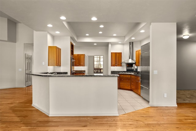 kitchen with light wood-style floors, a tray ceiling, wall chimney exhaust hood, a peninsula, and brown cabinetry