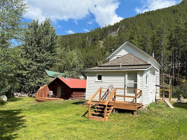 rear view of house with a lawn, roof with shingles, a deck, and a view of trees