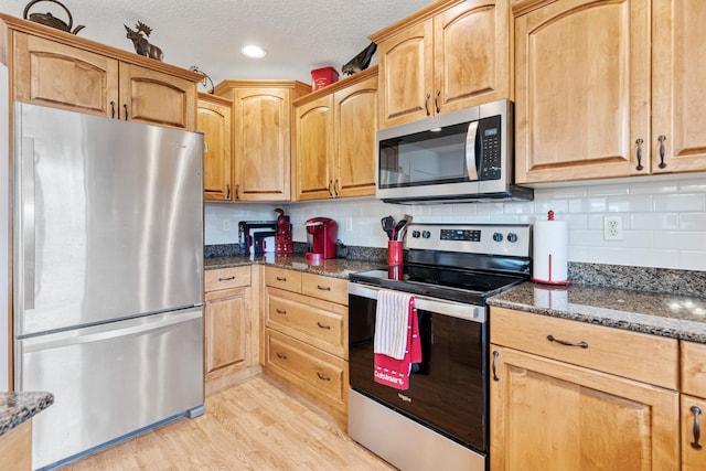 kitchen featuring light wood finished floors, stainless steel appliances, dark stone countertops, tasteful backsplash, and a textured ceiling