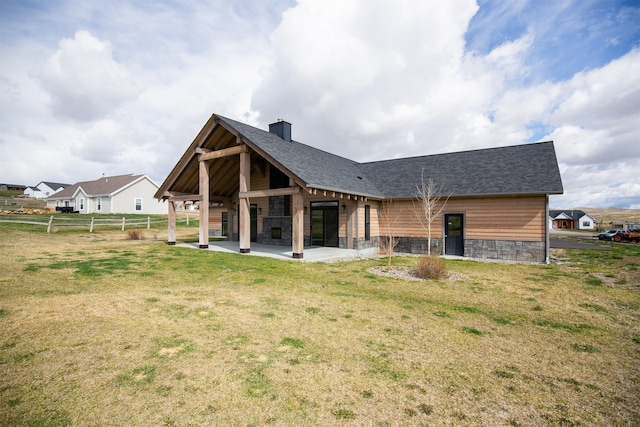back of property with a patio area, a lawn, stone siding, roof with shingles, and fence