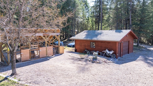 view of property exterior featuring a wooded view, a detached garage, a shingled roof, and an outbuilding