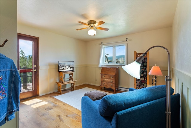 living area with wood finished floors, a ceiling fan, a wainscoted wall, and baseboard heating