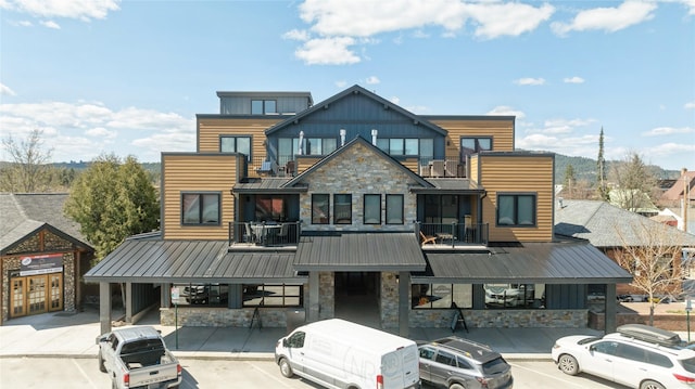view of front facade featuring board and batten siding, metal roof, uncovered parking, a balcony, and stone siding