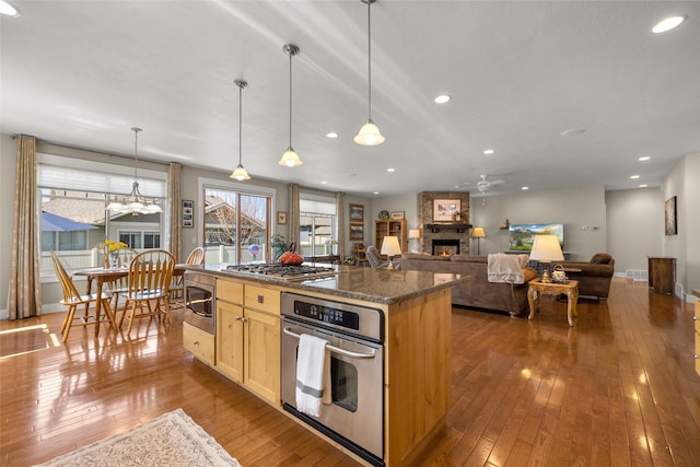 kitchen with ceiling fan, a stone fireplace, appliances with stainless steel finishes, light brown cabinets, and hardwood / wood-style floors