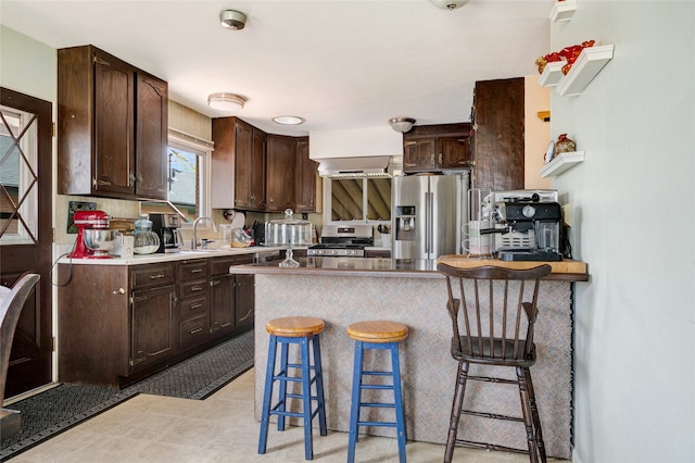 kitchen featuring wall chimney range hood, a kitchen breakfast bar, stainless steel appliances, and dark brown cabinets