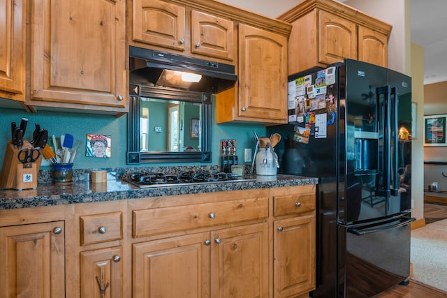 kitchen featuring stainless steel gas cooktop, dark countertops, under cabinet range hood, brown cabinets, and black fridge