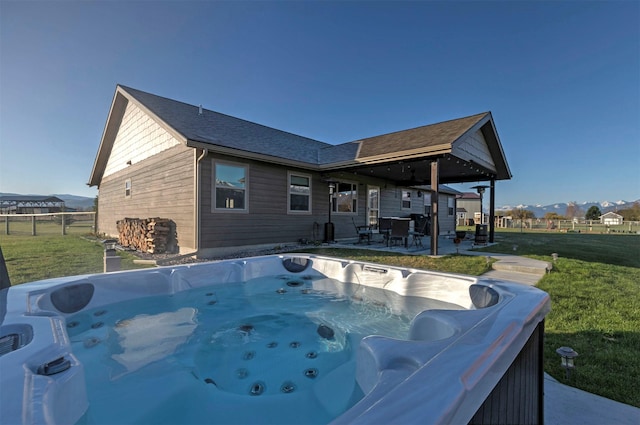 rear view of property featuring fence, a patio, a hot tub, a mountain view, and a yard