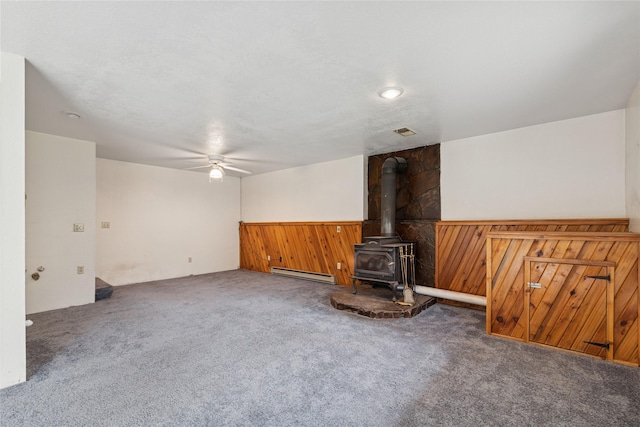 unfurnished living room with ceiling fan, a wood stove, carpet floors, a baseboard heating unit, and wooden walls