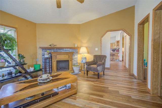 living room featuring ceiling fan, arched walkways, a fireplace with raised hearth, baseboards, and wood finished floors