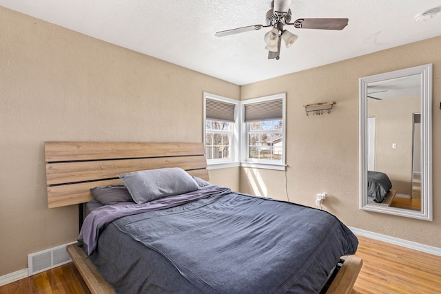 bedroom with visible vents, a textured wall, wood finished floors, baseboards, and a ceiling fan