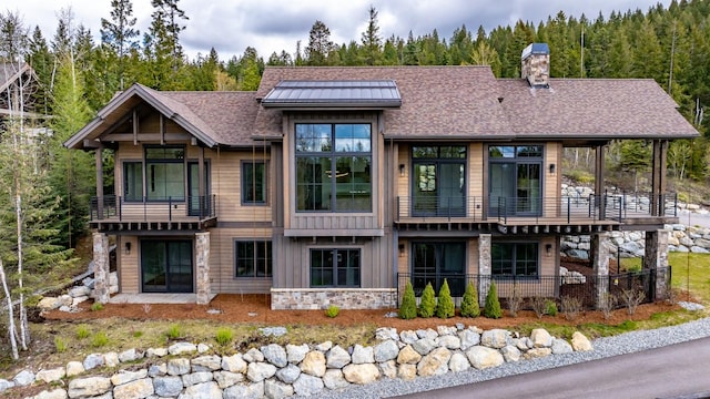 back of property with a forest view, a chimney, roof with shingles, a balcony, and stone siding