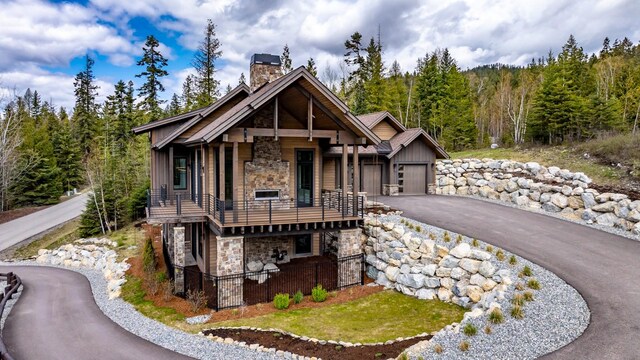 view of front of home with aphalt driveway, a balcony, stone siding, a chimney, and a forest view