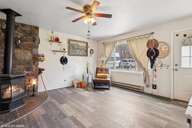 living area featuring wood finished floors, a wood stove, ceiling fan, and a baseboard heating unit