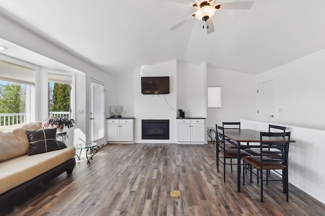 dining room featuring a ceiling fan, a glass covered fireplace, vaulted ceiling, and dark wood-style floors