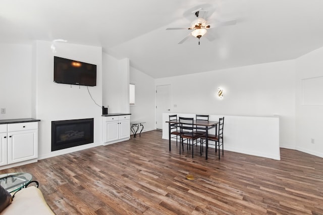 dining area with ceiling fan, vaulted ceiling, baseboards, dark wood-style floors, and a glass covered fireplace