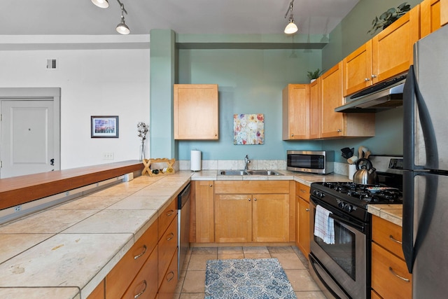 kitchen with track lighting, tile counters, under cabinet range hood, a sink, and appliances with stainless steel finishes