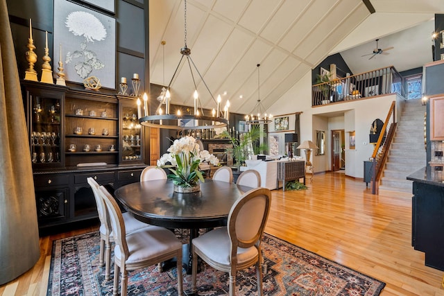 dining space with high vaulted ceiling, light wood finished floors, ceiling fan with notable chandelier, and stairs