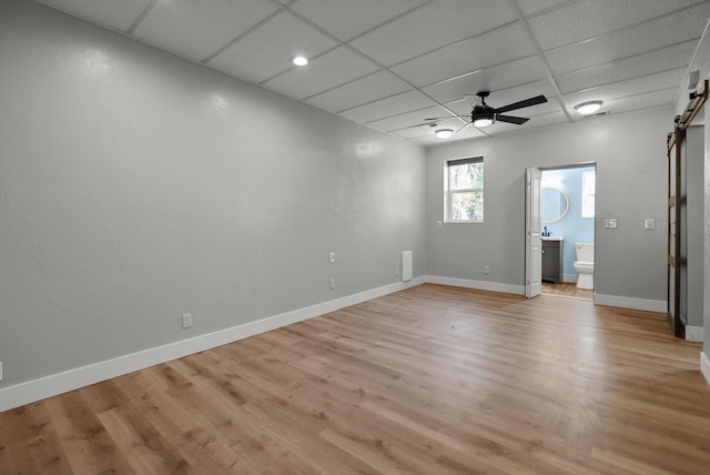 empty room featuring a ceiling fan, a barn door, baseboards, a drop ceiling, and light wood-style flooring