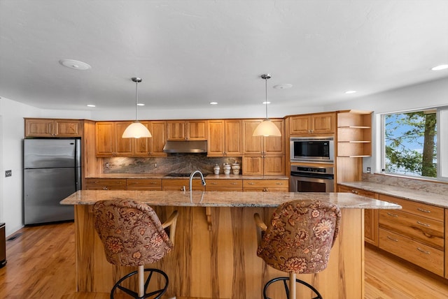 kitchen with light wood finished floors, open shelves, brown cabinetry, under cabinet range hood, and appliances with stainless steel finishes