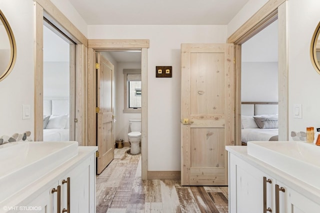 ensuite bathroom featuring toilet, ensuite bath, a sink, two vanities, and wood finished floors