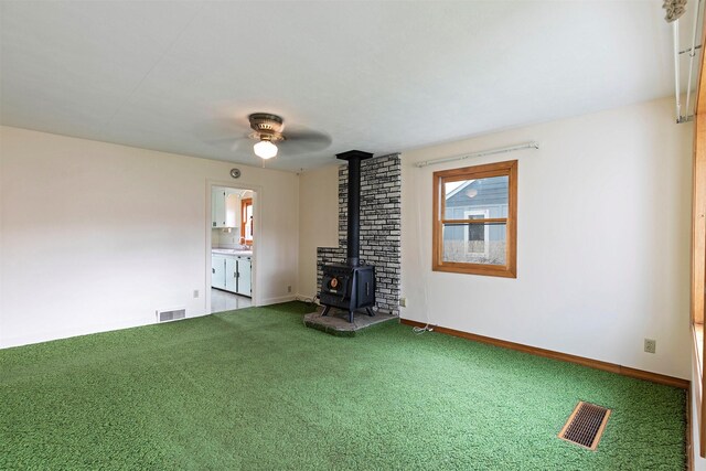 unfurnished living room with a wood stove, carpet flooring, visible vents, and ceiling fan