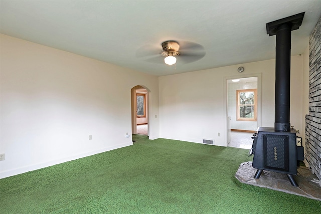 carpeted empty room featuring a wood stove, visible vents, arched walkways, a ceiling fan, and baseboards