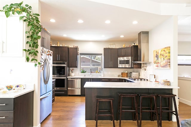 kitchen featuring a peninsula, dark brown cabinets, wall chimney exhaust hood, stainless steel appliances, and a kitchen breakfast bar