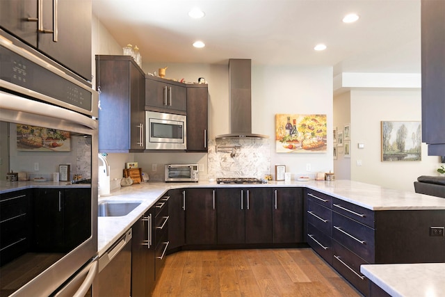 kitchen with appliances with stainless steel finishes, dark brown cabinets, a peninsula, wall chimney exhaust hood, and light wood-type flooring