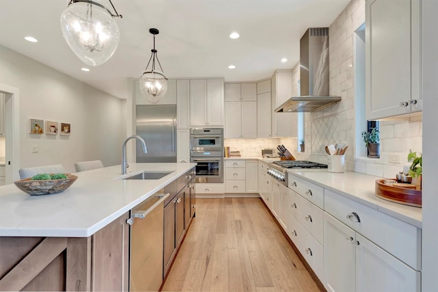 kitchen with a sink, wall chimney exhaust hood, a chandelier, light wood-style flooring, and appliances with stainless steel finishes