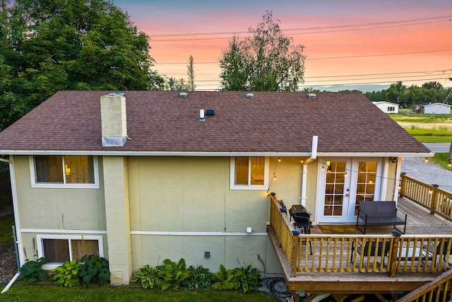 back of property at dusk featuring a shingled roof, stucco siding, french doors, and a deck