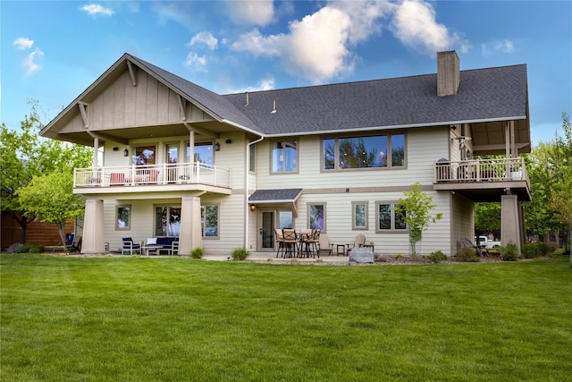 rear view of property with a lawn, a shingled roof, a chimney, and a balcony