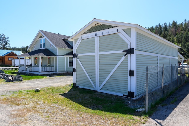 view of property exterior with covered porch, fence, a shingled roof, an outbuilding, and dirt driveway
