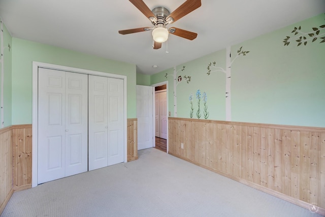 unfurnished bedroom featuring a wainscoted wall, wood walls, a closet, carpet flooring, and a ceiling fan