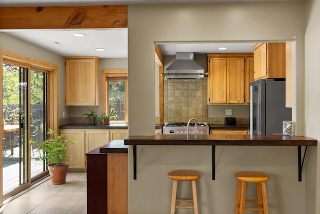 kitchen with wall chimney exhaust hood, freestanding refrigerator, dark countertops, decorative backsplash, and a breakfast bar area