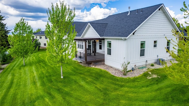 rear view of property featuring board and batten siding, roof with shingles, a deck, a lawn, and cooling unit