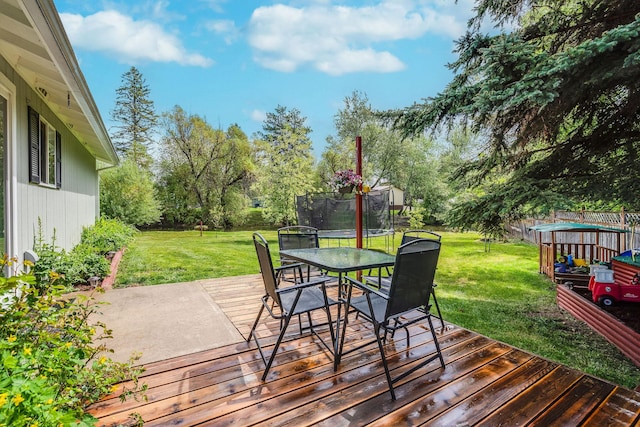 wooden deck with a trampoline, a lawn, outdoor dining area, and view of wooded area