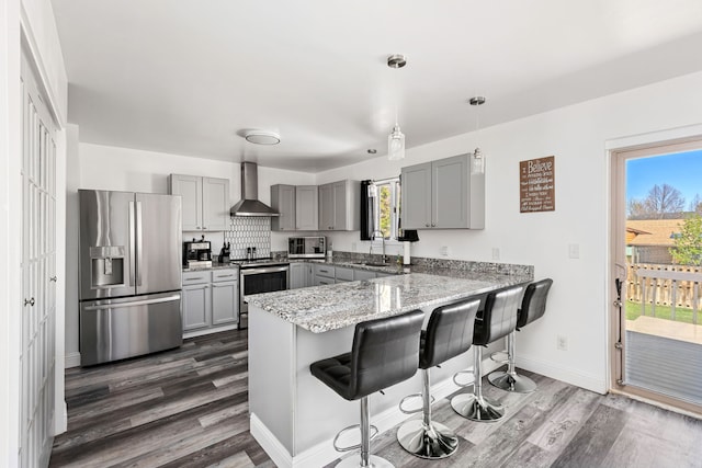 kitchen with appliances with stainless steel finishes, gray cabinets, wall chimney exhaust hood, a peninsula, and dark wood-type flooring