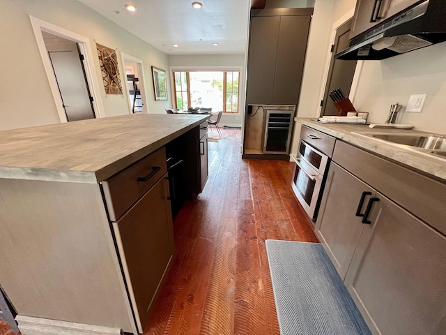 kitchen featuring under cabinet range hood, oven, wooden counters, beverage cooler, and dark wood-type flooring