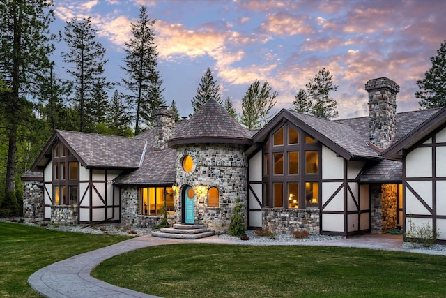 215 Browns Gulch Rd, Kalispell, MT 59901 - back of property at dusk with stone siding, stucco siding, a lawn, a chimney, and a high end roof