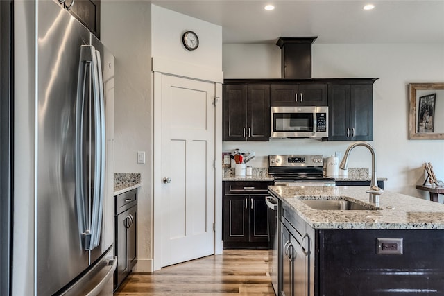 kitchen with stainless steel appliances, a sink, light wood-type flooring, an island with sink, and light stone counters