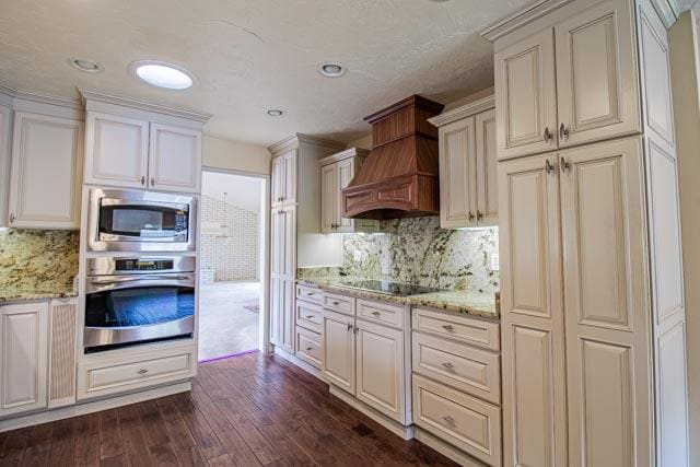 6 Cedar Lake Dr, Butte, MT 59701 - kitchen featuring appliances with stainless steel finishes, custom range hood, decorative backsplash, dark wood-type flooring, and light stone counters