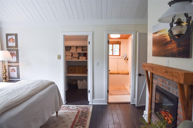 bedroom with dark wood finished floors, wood ceiling, baseboards, a brick fireplace, and ensuite bath