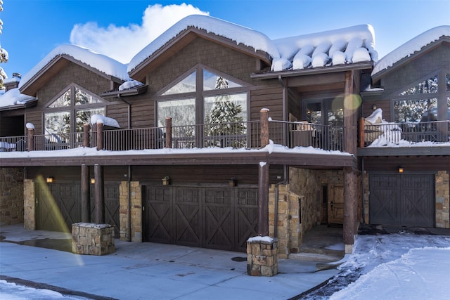 snow covered back of property with a garage, stone siding, and driveway