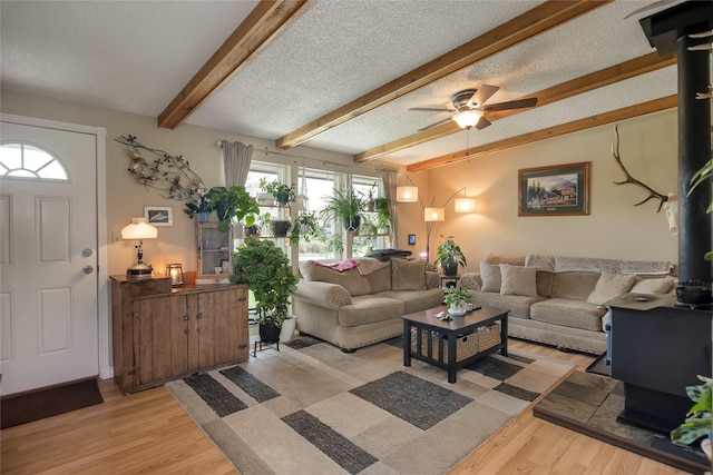 living area featuring light wood-type flooring, beamed ceiling, a ceiling fan, a wood stove, and a textured ceiling