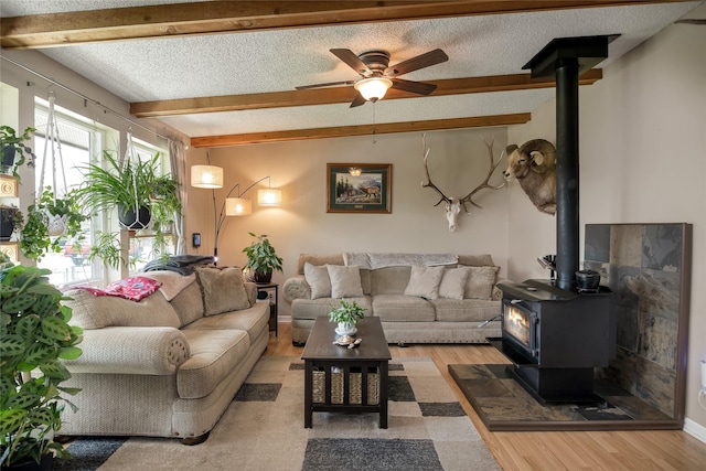 living room with a wood stove, beam ceiling, light wood-style floors, a textured ceiling, and a ceiling fan