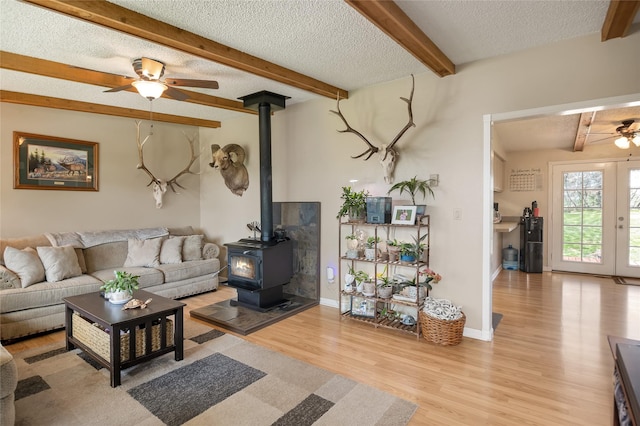 living room with a ceiling fan, a wood stove, wood finished floors, a textured ceiling, and beam ceiling