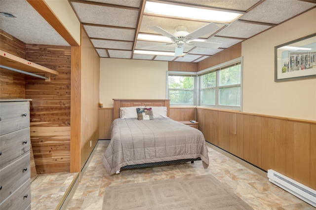 bedroom featuring wooden walls, a baseboard heating unit, a wainscoted wall, light stone finish floors, and a ceiling fan