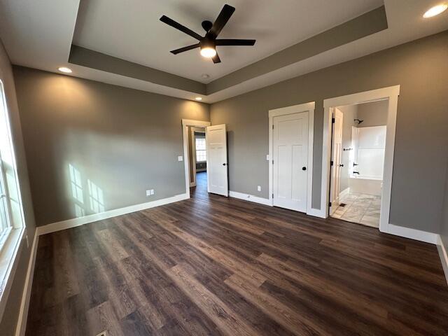 unfurnished bedroom with ensuite bathroom, a raised ceiling, ceiling fan, and dark wood-type flooring