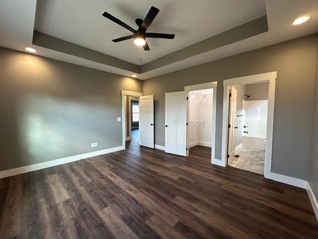 unfurnished bedroom featuring a walk in closet, ensuite bath, a raised ceiling, ceiling fan, and dark hardwood / wood-style floors