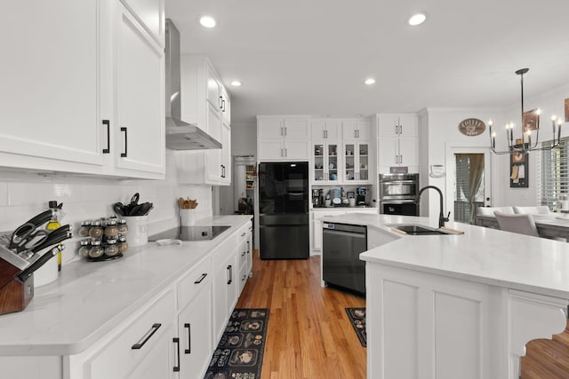 kitchen with wall chimney range hood, black appliances, sink, and a center island with sink